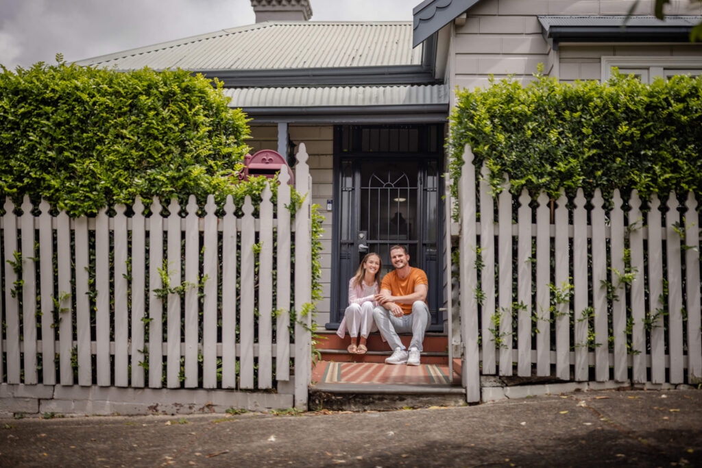 Young couple sitting, smiling, on the front step out the front of a single storey cottage home, who may have bought a home with the Help to Buy Scheme