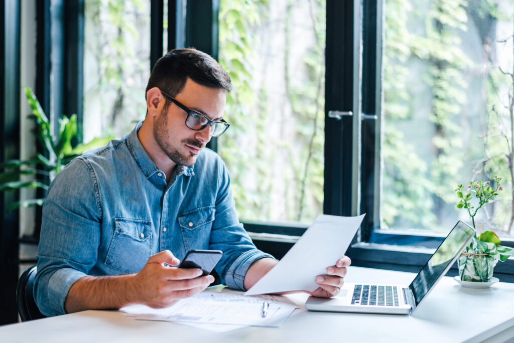 Man wearing glasses working at desk examining piece of paper, using phone, with laptop open